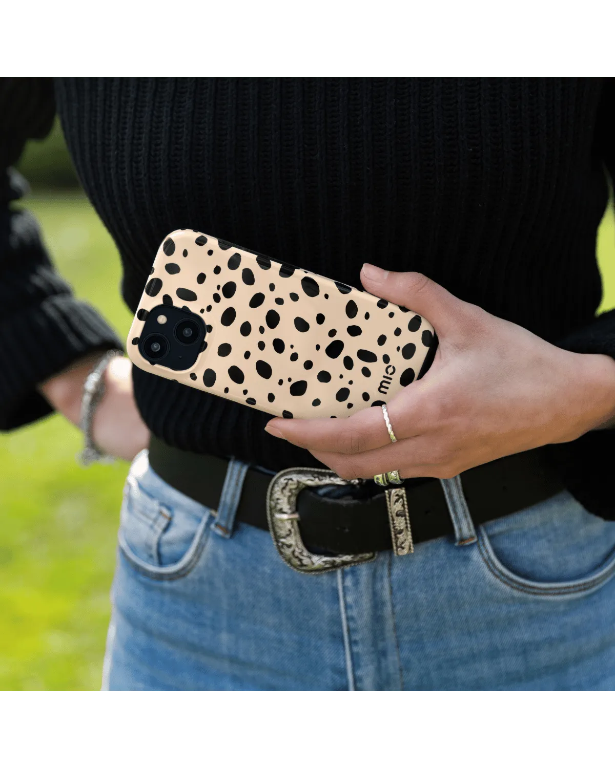 Hand houdt Samsung A55 5G met Mio hoes in zwart-bruine print, buiten in een natuurlijke omgeving.