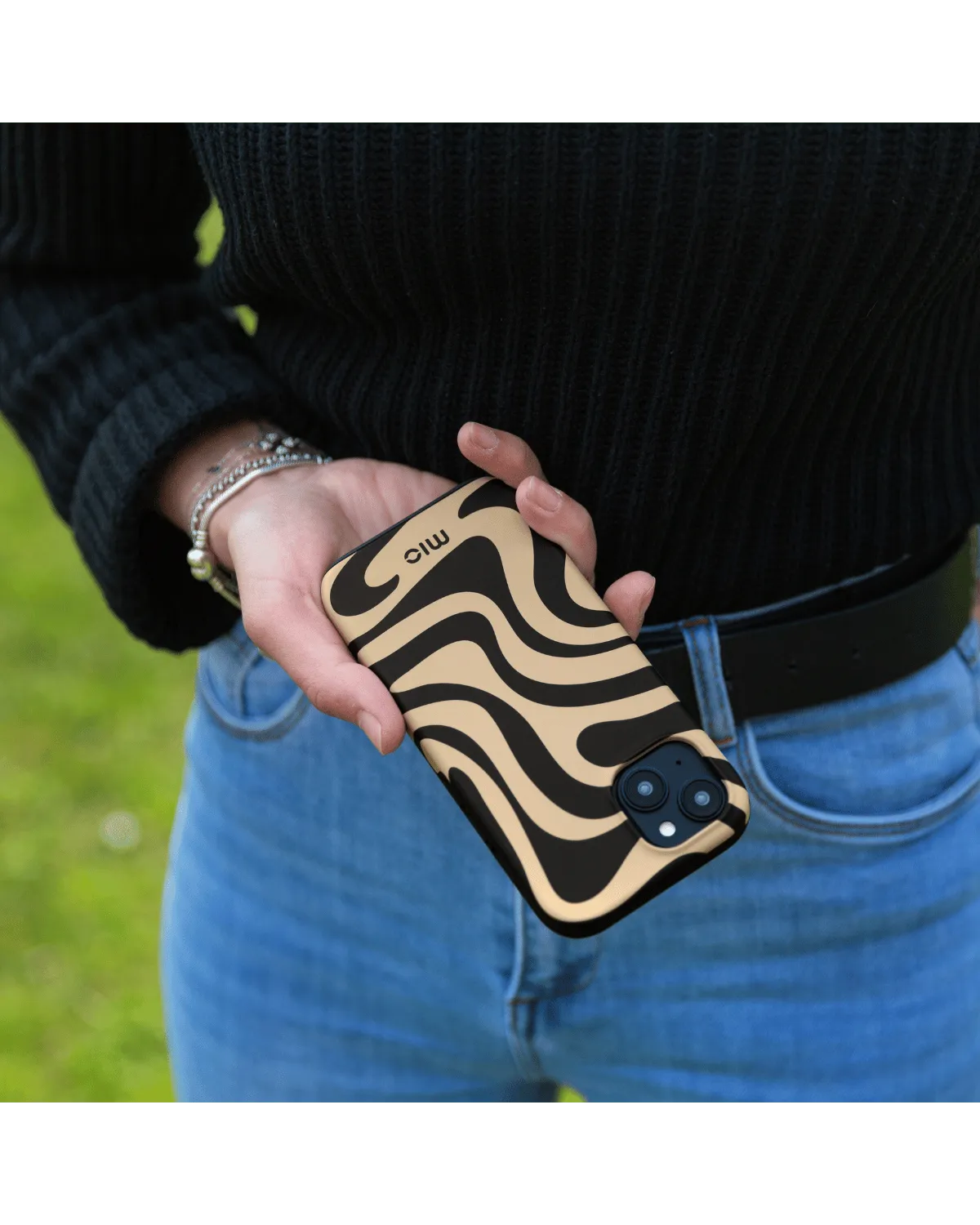 Persoon houdt zwart-bruin swirl hoesje van Mio by Mobilize vast, met Samsung Galaxy A55, in natuurlijke omgeving.